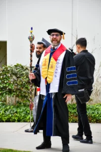 A person in academic regalia holds a ceremonial staff and stands outdoors near two other people. The person in focus wears a mortarboard, gown, and several honor stoles.