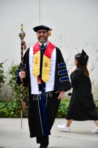 A person in academic regalia holds a ceremonial staff, wearing a black gown, colorful stole, and a cap. Another individual in a graduation cap and gown walks in the background.