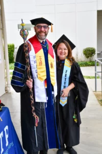 Two graduates in academic regalia pose for a photo; the left person holds a ceremonial staff, while the right person wears a sash labeled "FUTUR.