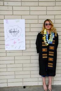 A person in academic regalia and leis stands by a white poster asking, "What kind of teacher do you want to be?" with various positive attributes written in colored text.