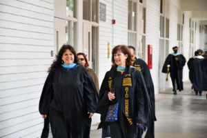 A group of people in academic regalia walk down a hallway. Two women in the foreground are smiling and talking.
