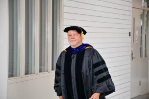 A person in academic regalia, including a cap and gown, stands in a hallway with white paneled walls, looking ahead.
