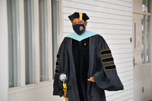 A person in academic regalia, including a cap and gown, stands holding a ceremonial mace. They are wearing a face mask and are in front of a white wall with windows and a door.