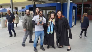 A group of people, including two in graduation gowns, pose for a photo outside a building. One person holds a camera, while others stand nearby.