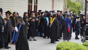 A group of people in academic regalia gather outdoors, conversing and preparing for a graduation ceremony.