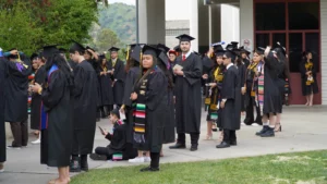 A group of graduates in caps and gowns stand outside, some looking at their phones, others talking. They all wear graduation stoles and are gathered near a building.