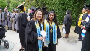 Two graduates in caps and gowns stand together, smiling, each wearing sashes labelled "TEACHER" and "FUTURE TEACHER." Others in graduation attire are in the background, some taking photos.