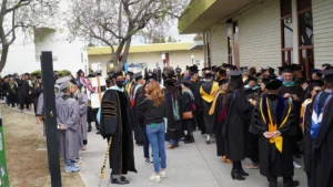 A group of graduates in caps and gowns gather outside a building, engaging in conversation and preparing for a ceremony. People are lined up, with some facing forward and others interacting.