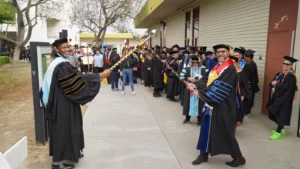 Professors in academic regalia holding ceremonial maces lead a procession of graduates dressed in caps and gowns outside a building during a graduation ceremony.
