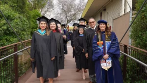 A group of graduates in academic regalia stand outdoors on a pathway, smiling. Some wear hoods and stoles, and one individual in front wears a blue doctoral gown with various sashes and cords.