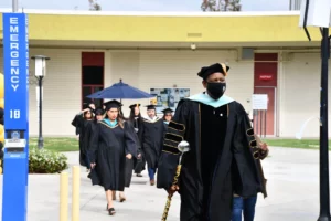 A masked academic leads a graduation procession in black gowns and caps, with one graduate holding an umbrella. An "EMERGENCY" station is visible on the left side of the image.