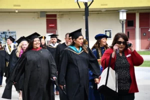 A group of graduates in caps and gowns walk outside. A woman in front is talking on the phone and carrying a bag.