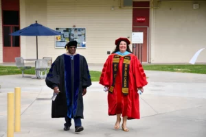 Two individuals wearing academic regalia walk outside a building. One wears black robes and the other wears a red doctoral gown and cap.
