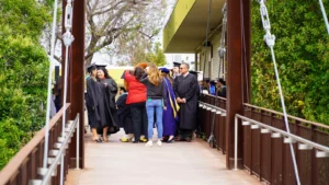 A group of people in graduation attire gather on a narrow bridge, some smiling and chatting, surrounded by greenery and a building in the background.