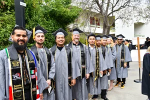 A line of graduates in caps and gowns stand outdoors, posing for a group photo. Some wear stoles representing different flags. Trees and a building are visible in the background.