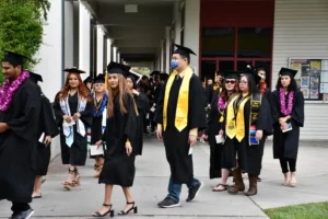 A group of students in graduation attire, including caps and gowns, walk down a corridor. Some wear leis and stoles, one student wears a face mask. They appear to be heading to or from a ceremony.