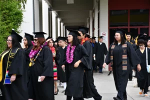 A group of graduates in black caps and gowns, some wearing leis and cords, walk together in an outdoor corridor, celebrating their graduation.