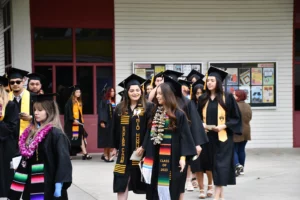 Graduates in caps and gowns, adorned with sashes and leis, walk together outside of a building. Some are smiling and talking, and others are in the background.