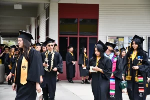 A group of graduates in caps and gowns walk in a procession outside a building. Some hold their diplomas and wear leis or sashes.
