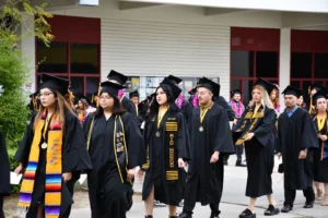 A group of graduates in caps and gowns walk in a line during a ceremony. Some wear honor cords and stoles, and many have medallions around their necks.