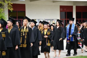 A group of graduates in caps and gowns, some wearing stoles, are walking in a line during a graduation ceremony.