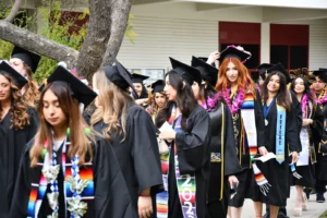 A group of graduates in caps and gowns walk in a line during a graduation ceremony. Some wear colorful sashes and flower leis.