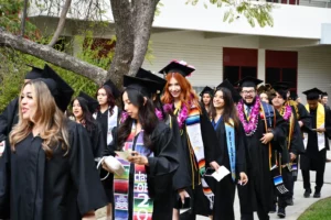 A group of graduates in caps and gowns walk in a procession outdoors, some wearing graduation stoles and leis, smiling and celebrating their achievements.