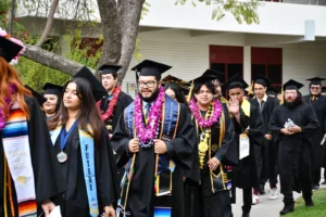 Graduates in caps and gowns walk in a procession. Some wear leis and cords. One woman has a "Future" sash, and another holds a rolled diploma. Trees and a building are in the background.