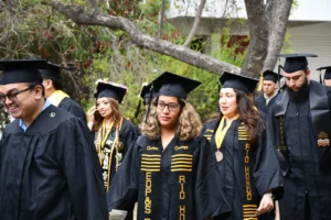 Graduates in caps and gowns walk in a line during an outdoor graduation ceremony. Trees and a building are in the background.