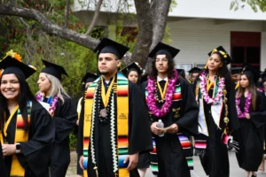 Graduates in caps and gowns walk in a procession. Some wear colorful sashes and leis. Trees and a building are in the background.