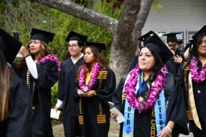 A group of graduates in caps and gowns, some wearing leis, walk outdoors. One graduate holds a phone, another holds a bottle and a ribboned item. Trees and a building are visible in the background.