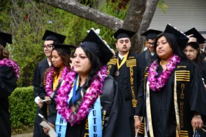 A group of graduates wearing black caps and gowns, adorned with floral leis, walk outdoors near a tree and greenery, celebrating their commencement. Some display honor stoles and medals.