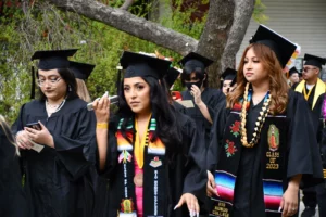 Group of students in graduation caps and gowns walking outside. One student in front is adjusting her tassel, while others follow behind her. Tree and greenery are visible in the background.