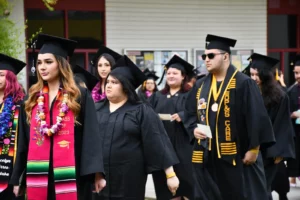 A group of graduates in caps and gowns, some wearing sashes and leis, stand together outdoors during a graduation ceremony.