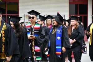 Graduates in caps and gowns, many wearing colorful stoles, walk in a procession during a graduation ceremony.