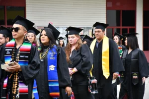 A group of graduates in black gowns and caps stand in line, some wearing colorful stoles and sashes. They appear to be at a graduation ceremony.