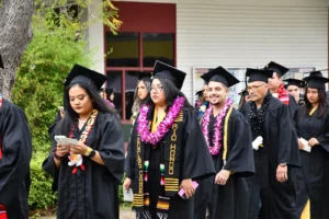A group of graduates in caps and gowns, adorned with leis and stoles, proceed in a line during a graduation ceremony, with one graduate looking at a phone.