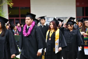 A group of graduates in black caps and gowns, adorned with various sashes and leis, walk in a procession during a graduation ceremony.