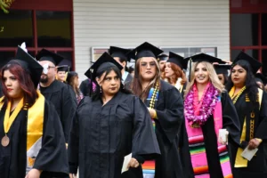 A diverse group of students in black graduation gowns and caps stands together outdoors. Some wear colorful stoles, leis, or honor cords. They appear to be at a graduation ceremony.