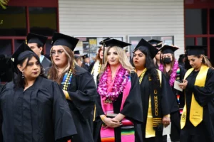 A group of graduates in black caps and gowns stand together, with one graduate in the center wearing a pink lei.