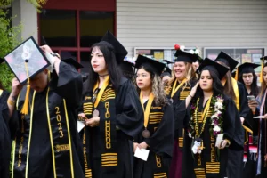 A group of graduates in black caps and gowns, adorned with yellow sashes, stand together. Some hold papers, and the front graduate's cap reads "2023" and "RISE.