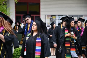A group of graduates in black caps and gowns, some wearing colorful stoles and leis, walk outside near a building with bulletin boards. Two graduates in the foreground have "Class of 2023" sashes.