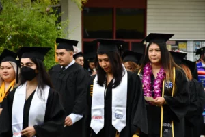 A group of graduates in black caps and gowns walk in a procession. Some are wearing stoles and leis. One graduate on the left is wearing a face mask.