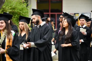 A group of graduates in black caps and gowns stands in line outdoors. Some are holding papers and looking around. A few graduates have yellow stoles. Trees and a building are in the background.