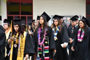 A group of graduates in caps and gowns, adorned with leis and stoles, walk together during a commencement ceremony outside a building.
