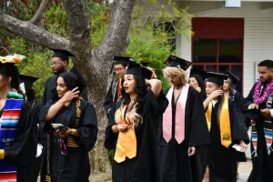 A group of graduates in caps and gowns walk together outdoors. Some adjust their attire while others interact with their surroundings. Trees and a building are visible in the background.