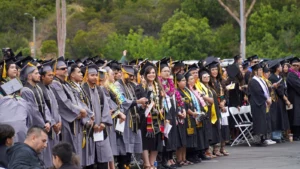 A large group of graduates in caps and gowns stand in rows during an outdoor graduation ceremony, with some holding programs and leis, surrounded by trees and seated attendees.