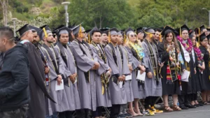 People in graduation gowns and caps standing in line outdoors, with some wearing leis and stoles, during a graduation ceremony. Trees and greenery are visible in the background.