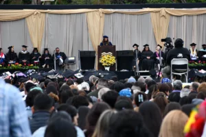 An individual speaks at a podium adorned with flowers during a graduation ceremony. Graduates in caps and gowns sit onstage, and a seated audience watches. A camera operator films the event.
