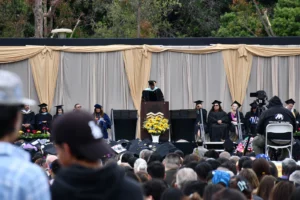 A commencement speaker addresses a crowd at a graduation ceremony. Be-robed graduates sit on stage, and audience members watch from seating below.